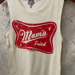 May include: White sleeveless tank top with a red distressed graphic that says "Mama Tried" with the words "Country Deep" underneath. The tank top has a tag that says "Country Deep" with a size "M" on it.