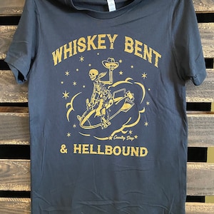 Könnte beinhalten: Schwarzes T-Shirt mit goldfarbener Grafik, die ein Skelett auf einer Bombe reitend, mit Cowboyhut und dem Text "Whiskey Bent & Hellbound" zeigt. Das T-Shirt hängt vor einem Holz Hintergrund.