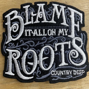 Può includere: Toppa ricamata nera con cuciture bianche. La toppa dice "Blame it all on my roots" con un fiocco decorativo e "Country Deep" in basso.