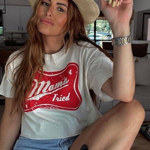 Op de afbeelding: Een vrouw draagt een wit T-shirt met een rode en witte afbeelding waarop "Mama's Tried" staat. Ze draagt ook een strooien cowboyhoed, een spijkerbroekje en een horloge.