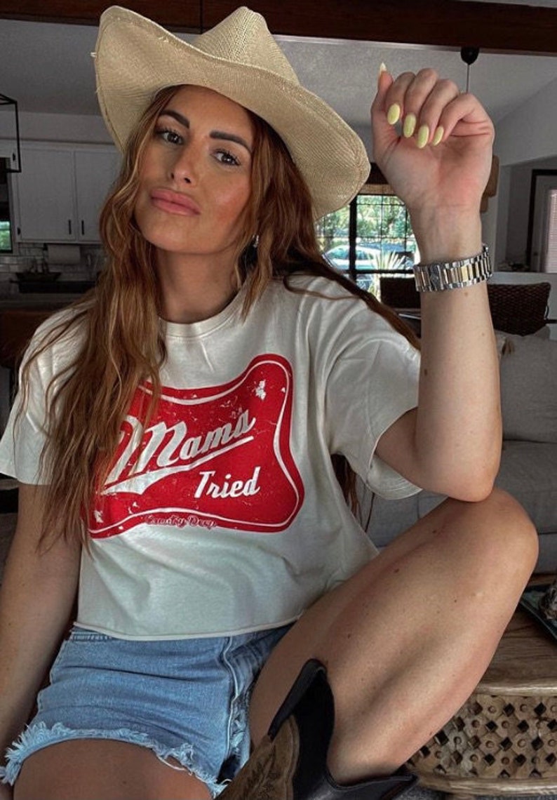 Op de afbeelding: Een vrouw draagt een wit T-shirt met een rode en witte afbeelding waarop "Mama's Tried" staat. Ze draagt ook een strooien cowboyhoed, een spijkerbroekje en een horloge.