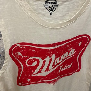 May include: A white sleeveless tank top with a red distressed graphic of a beer bottle label that says "Mama Tried" and "Country Deep".