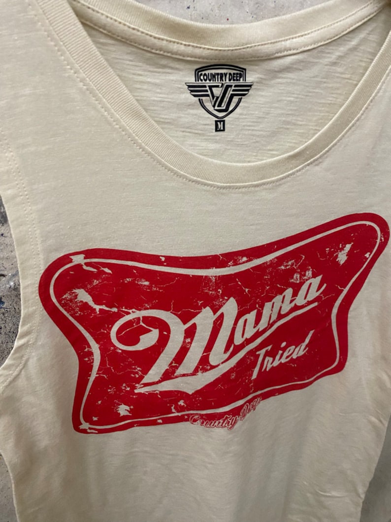May include: A white sleeveless tank top with a red distressed graphic of a beer bottle label that says "Mama Tried" and "Country Deep".