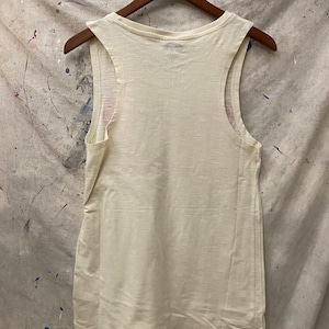 May include: A white sleeveless tank top with a scoop neck. The tank top is hanging on a wooden hanger against a textured background.