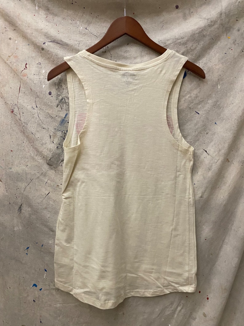 May include: A white sleeveless tank top with a scoop neck. The tank top is hanging on a wooden hanger against a textured background.