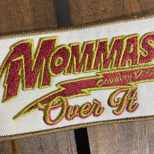 May include: A white embroidered patch with the text "Momma's Country Delights" in red and gold lettering. The text "Over It" is also embroidered in gold lettering.