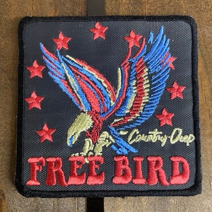 May include: A black patch with a red, white, and blue embroidered eagle with stars around it. The text "Free Bird" is embroidered in red below the eagle. The text "Country Cheap" is embroidered in blue in the bottom right corner.