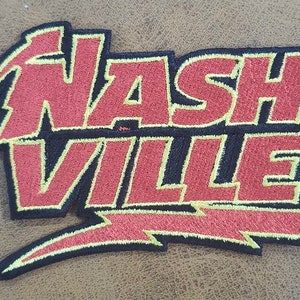 Op de afbeelding: Geborduurde patch met het woord "NASHVILLE" in rood, omlijnd met zwart en geel. Het ontwerp bevat een bliksemschicht en gestileerde letters, ideaal voor jassen, tassen of andere textielartikelen.