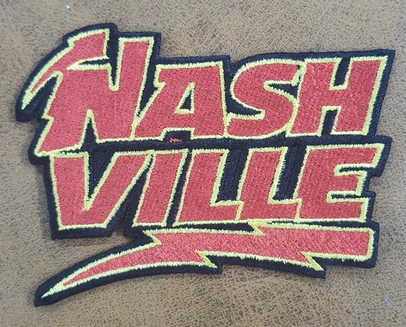 May include: Embroidered patch with the word "NASHVILLE" in red, with black and yellow outlines. The design incorporates a lightning bolt and stylized lettering, suitable for application on clothing or accessories.
