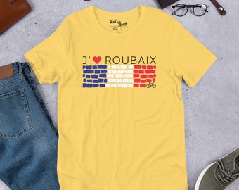 May include: A yellow t-shirt featuring the text "J'❤️ ROUBAIX" above a graphic of the French flag made of bricks and a bicycle. The shirt has a crew neck and short sleeves. The text "Hell of the North" is printed on the upper part of the shirt.