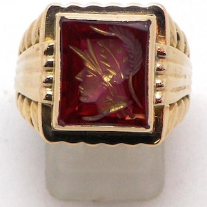 Puede incluir: Un anillo dorado con una piedra rectangular de color rojo intenso. La piedra presenta un perfil detallado de una figura con casco y corona de laurel. El anillo tiene un diseño de banda acanalada y una estética clásica y vintage.