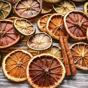 Puede incluir: Una colección de rodajas de cítricos secos en tonos naranja, marrón y verde, junto con dos palitos de canela. Las rodajas están dispuestas sobre una superficie de madera clara, creando una estética rústica y natural.