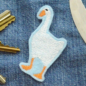 May include: Embroidered goose patch with a light blue outline and body, white center, and orange beak and feet. The patch is on a denim background, with keys and a knife visible.