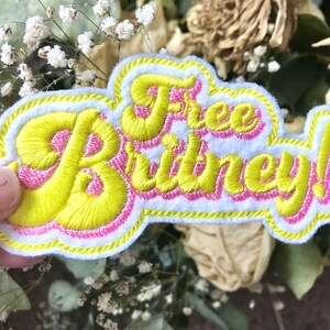 May include: Embroidered patch with the text "Free Britney!" in yellow script with a pink outline. The patch has a white background and a yellow and white border. The patch is a statement piece.