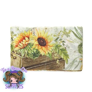 May include: A roll of ribbon featuring a sunflower design. The ribbon displays two large sunflowers with yellow petals and brown centers, set against a wooden crate and green foliage. The background is a light, textured pattern.