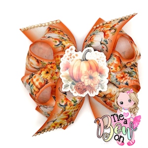 May include: A large, decorative hair bow featuring a pumpkin and floral design. The bow is primarily orange with a repeating pattern of pumpkins and flowers. The bow has a rhinestone trim and a central pumpkin sticker. The text "Tie a Bow on" is visible.