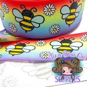 May include: Rainbow-hued ribbon with a repeating pattern of cartoon bees and white daisies. The ribbon has the text "Tie a Bow On" and the word "SUPPLIES" in the image.