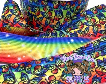 May include: A roll of vibrant ribbon with a butterfly design in red, yellow, blue, and green. A rainbow-coloured ribbon is also visible. The text "Tie a Bow On SUPPLIES" is printed on the ribbon.