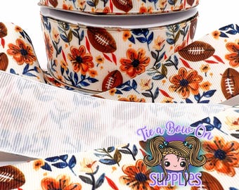 May include: Cream-colored ribbon with a repeating pattern of brown footballs and orange flowers with blue leaves. The ribbon is stacked in rolls, with some unrolled ribbon in the foreground. The text "Tie a Bow On SUPPLIES" is visible.