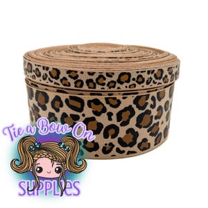 May include: A spool of tan ribbon with a leopard print design. The ribbon is wrapped around itself in a circular shape. The text "Tie a Bow On SUPPLIES" is in the lower left corner.