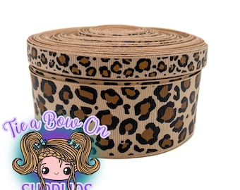 Può includere: Una bobina di nastro beige con stampa leopardata. Il nastro è avvolto a cerchio. Il testo "Tie a Bow On SUPPLIES" è visibile nell'angolo in basso a sinistra.