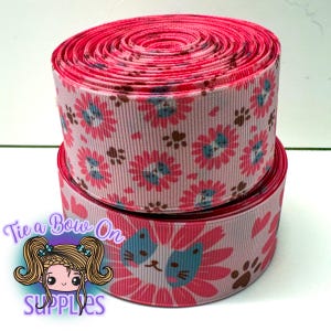 May include: Two rolls of pink ribbon with a floral and paw print design. The ribbon displays pink flowers with blue centres and brown paw prints on a light pink backdrop. The top roll is tightly wound, while the bottom roll shows the pattern more clearly.