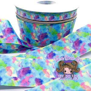 May include: A spool of grosgrain ribbon featuring a vibrant watercolor-style print with blended patches of blue, green, pink, and purple. The ribbon is wrapped around a white plastic core and includes a logo with the text Tie a Bow On Supplies.