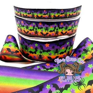 Könnte beinhalten: Ein Stapel aus drei Ripsbandrollen mit einem sich wiederholenden Muster aus schwarzen Spinnen, Fledermäusen und bunten Sternen auf einem leuchtenden Regenbogen-Verlaufshintergrund. Der Text Tie a Bow On SUPPLIES steht unten rechts.