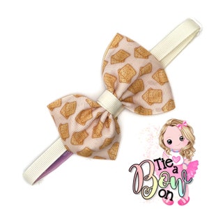 May include: A white bow with a brown and white cracker pattern. The bow is attached to a white elastic headband.