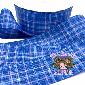May include: Blue plaid ribbon with white and light purple accents. The ribbon is curled and folded, with the text "Tie a Bow On SUPPLIES" visible in the lower right corner. This ribbon is suitable for crafting and gift wrapping.