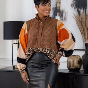 May include: A long-sleeved blouse featuring a brown and black checkered bodice with a ruffled collar, paired with sleeves displaying large orange, beige, and black circular patterns. The hem is accented with a leopard print band and cuffs.