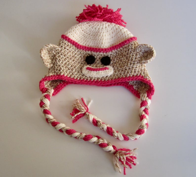 Pu&ograve; includere: Un cappello da scimmia all'uncinetto con un pompon rosa in cima, orecchie marroni e un cordoncino intrecciato rosa e bianco.