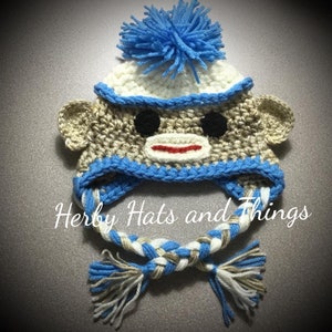 Pu&ograve; includere: Cappello da scimmia all'uncinetto con strisce blu e bianche, un pon pon blu e una cravatta intrecciata marrone e bianca.