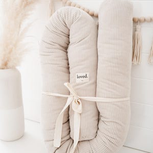 Könnte beinhalten: Ein beiges U-förmiges Kissen aus Cord, gebunden mit einem cremefarbenen Band. Das Kissen hat ein Etikett mit der Aufschrift "loved". Der Hintergrund umfasst eine weiße Wand, eine Vase mit Pampasgras und eine Holzperlen-Girlande mit Quasten.