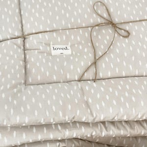 Könnte beinhalten: Eine beige gesteppte Baby-Spielmatte mit einem weißen Blatt-Muster. Die Matte ist mit Schnur gebunden und hat ein Etikett mit der Aufschrift "loved."