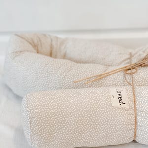 Könnte beinhalten: Ein Baby-Nest und ein Stützkissen mit beigem und weißem Muster. Der Stoff hat ein zartes blattartiges Muster. Das Kissen ist mit Bindfaden gebunden und trägt das Etikett "loved."