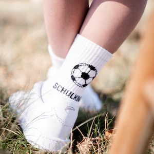 Könnte beinhalten: Weiße Socken mit einem schwarzen Fußball-Design und dem Wort "SCHULKIN" an der Seite. Die Socken werden an den Füßen einer Person getragen.