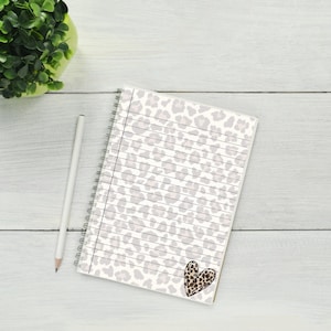Pode incluir: Um caderno espiral com fundo branco e um padrão de estampa de leopardo rosa claro e marrom. O caderno tem páginas pautadas e um pequeno coração marrom com um design de estampa de leopardo no canto inferior direito.