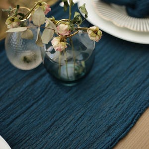May include: A table setting featuring a textured, dark teal runner. Two small glass vases with dried flowers are in the foreground. White plates with a decorative edge and a teal napkin are in the background. The scene evokes a rustic, elegant aesthetic.
