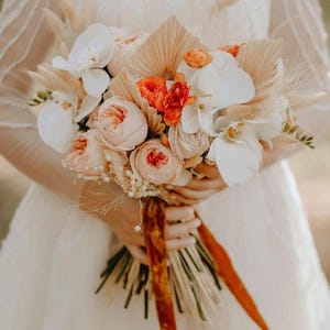 May include: A bridal bouquet with peach and white flowers, orange flowers, and dried grasses tied with a rust-colored ribbon.
