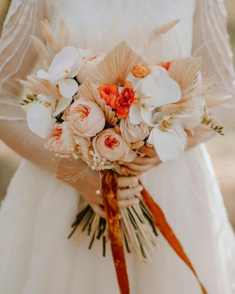 May include: A bridal bouquet with peach and white flowers, orange flowers, and dried grasses tied with a rust-colored ribbon.