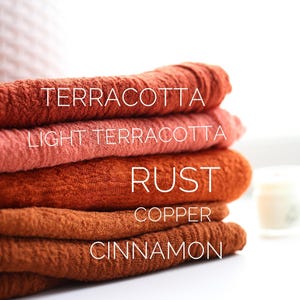May include: A stack of five folded textiles in shades of terracotta, light terracotta, rust, copper, and cinnamon. The names of the colors are written on the textiles in white. A white candle is in the background.