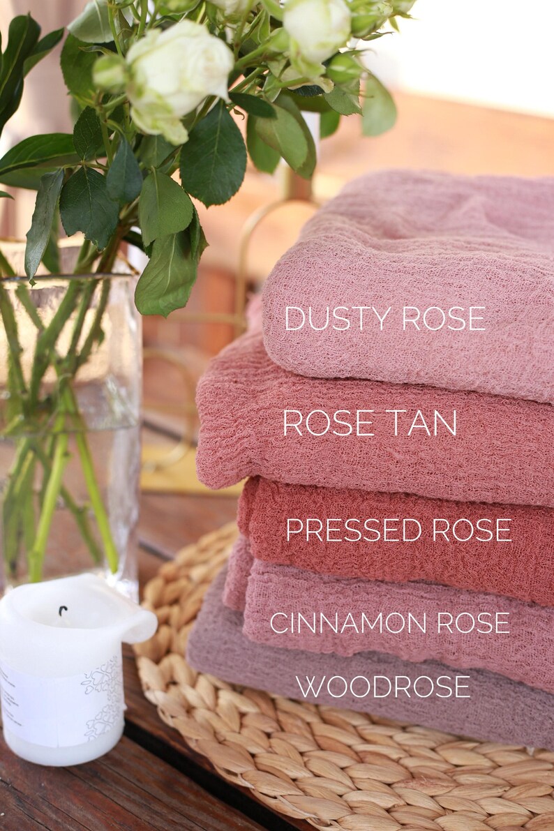 a stack of pink towels with labels for each color, including "DUSTY ROSE", "ROSE TAN", "PRESSED ROSE", "CINNAMON ROSE", and "WOODROSE". There is also a vase with white flowers and a candle on a woven placemat, creating a cozy and relaxing atmosphere.