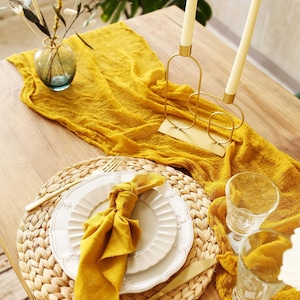 May include: A table setting with a yellow runner, a woven placemat, and white plates. A gold candle holder with two white candles and a small blue vase with greenery are also present. The overall theme is a warm, inviting dining arrangement.
