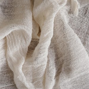 May include: A piece of off-white fabric with a loose, gauzy weave and a crinkled, textured surface. The material is draped in soft folds, showcasing its semi-sheer quality and the irregular, open-knit pattern throughout the entire textile.