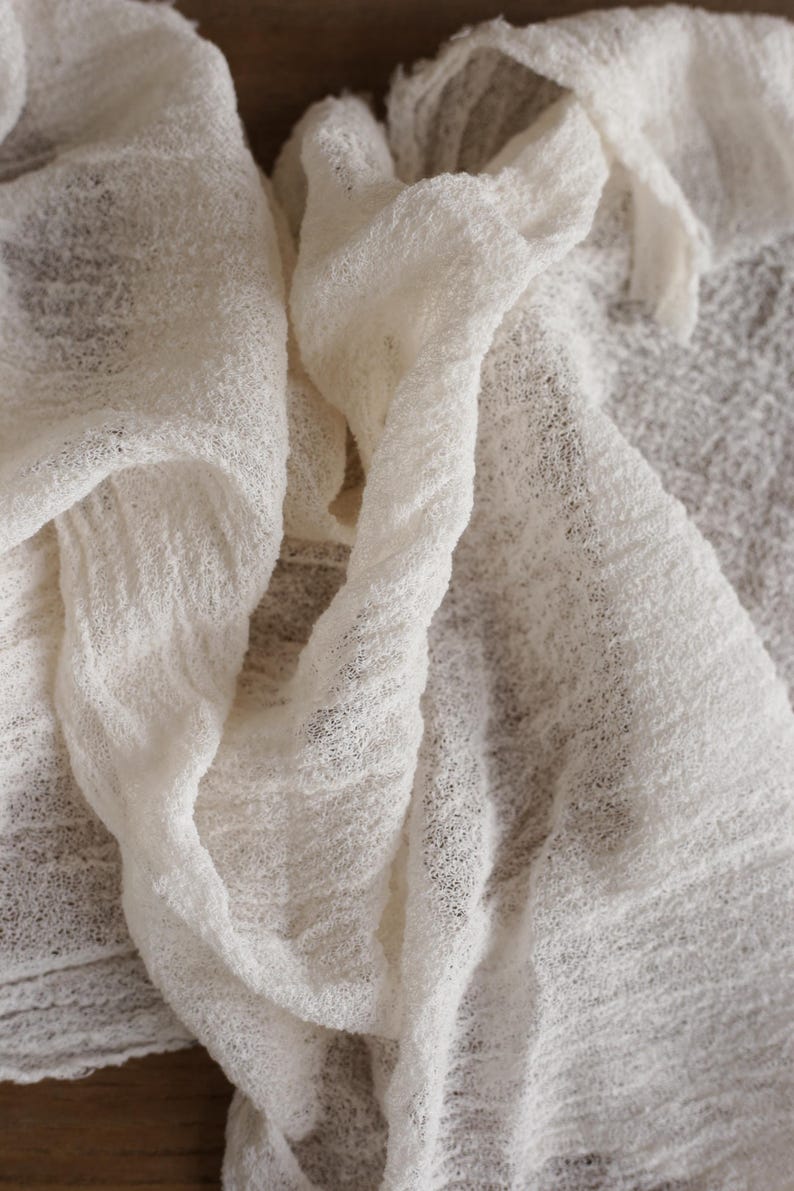 May include: A piece of off-white fabric with a loose, gauzy weave and a crinkled, textured surface. The material is draped in soft folds, showcasing its semi-sheer quality and the irregular, open-knit pattern throughout the entire textile.