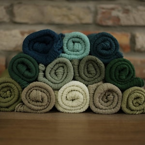 Puede incluir: Una pila de tela enrollada en varios tonos de verde, turquesa y beige. Los rollos están cuidadosamente dispuestos, mostrando una gama de colores que van desde el verde oscuro del bosque hasta el verde salvia claro. La tela parece suave.