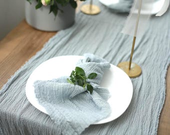 Könnte beinhalten: Ein staubblauer Tischläufer aus Mullstoff mit knittriger Textur und ausgefransten Kanten. Eine passende staubblaue Serviette liegt gefaltet auf einem weißen Teller, verziert mit einem kleinen grünen Zweig. Der Läufer liegt auf einem Holztisch.