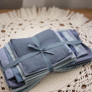 Op de afbeelding: Een stapel blauwe stoffen vierkanten, vastgebonden met een bijpassend lint. De stof heeft een zachte, getextureerde uitstraling, met verschillende tinten blauw. De bundel ligt op een crèmekleurig, gehaakt kleedje.