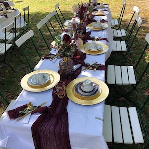 May include: A long table set for an outdoor event, featuring white tablecloths, gold chargers, and elegant china. Burgundy runners and floral arrangements add color. White folding chairs with green frames are arranged around the table, set on a grassy lawn.